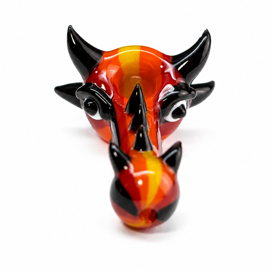 A hand-blown glass sculpture of a horned demon or beast head, featuring a glossy finish with vibrant orange, red, and yellow flame-like patterns and contrasting black horns and facial details, set against a plain white background.